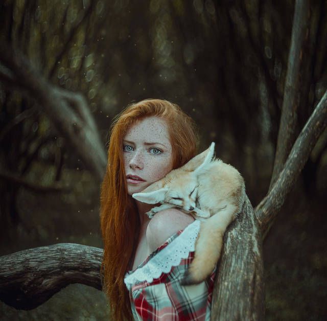 a young woman with long red hair and freckles stands in a forest, a fennec fox nestled peacefully in the crook of her arm. the woman gazes directly at the viewer with a serene expression, her shoulder bare and adorned with a white lacetrimmed garment. the scene is bathed in soft, diffused natural light, creating an ethereal and magical atmosphere. the color palette consists mainly of earthy browns, greens, and muted blues.  the image evokes a sense of connection and harmony between the woman and the wild animal.