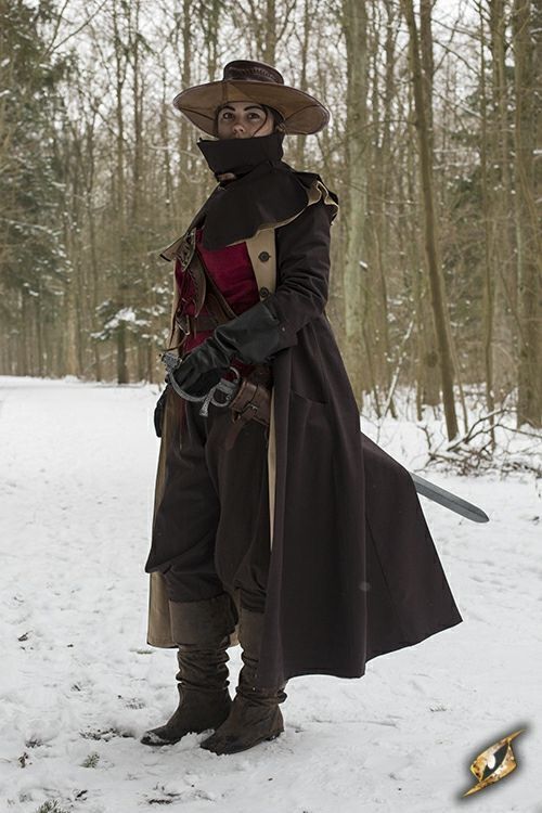 a woman in a dark coat, hat and boots with a sword strapped to her side and a cloak standing in a snowy forest.