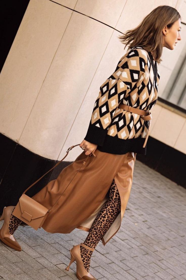 a stylish woman is captured in a fulllength shot, showcasing her autumnal outfit. she's wearing a black and beige geometric patterned cardigan, a brown leather midi skirt with a thighhigh slit, and leopard print tights. her accessories include brown pointedtoe high heels and a brown crossbody bag. the photo is taken outdoors against a light beige wall, adding a contrast to her attire. the overall style is chic and trendy, with the model posing effortlessly.