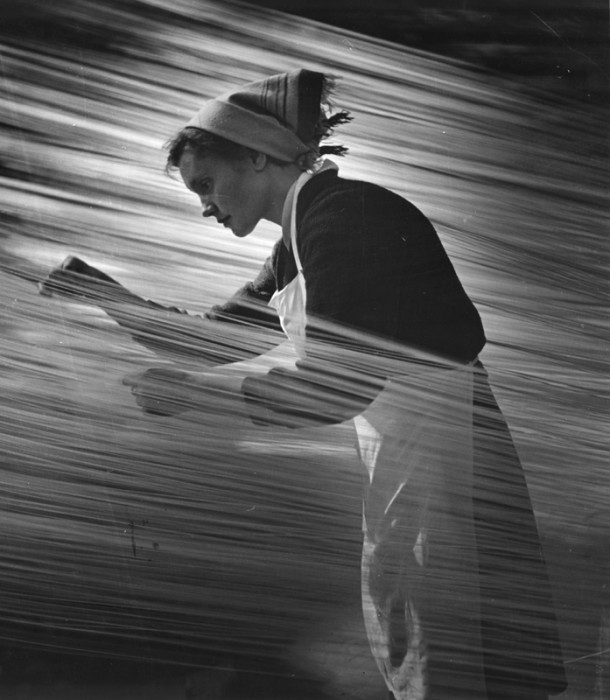 a black and white photograph of a woman working in a factory. the image is taken from the side and the woman is facing left. she is wearing a headscarf, a dark sweater, and a white apron. her hands are blurred, suggesting she is working with something in motion. the background is also blurred, creating a sense of movement and industry.