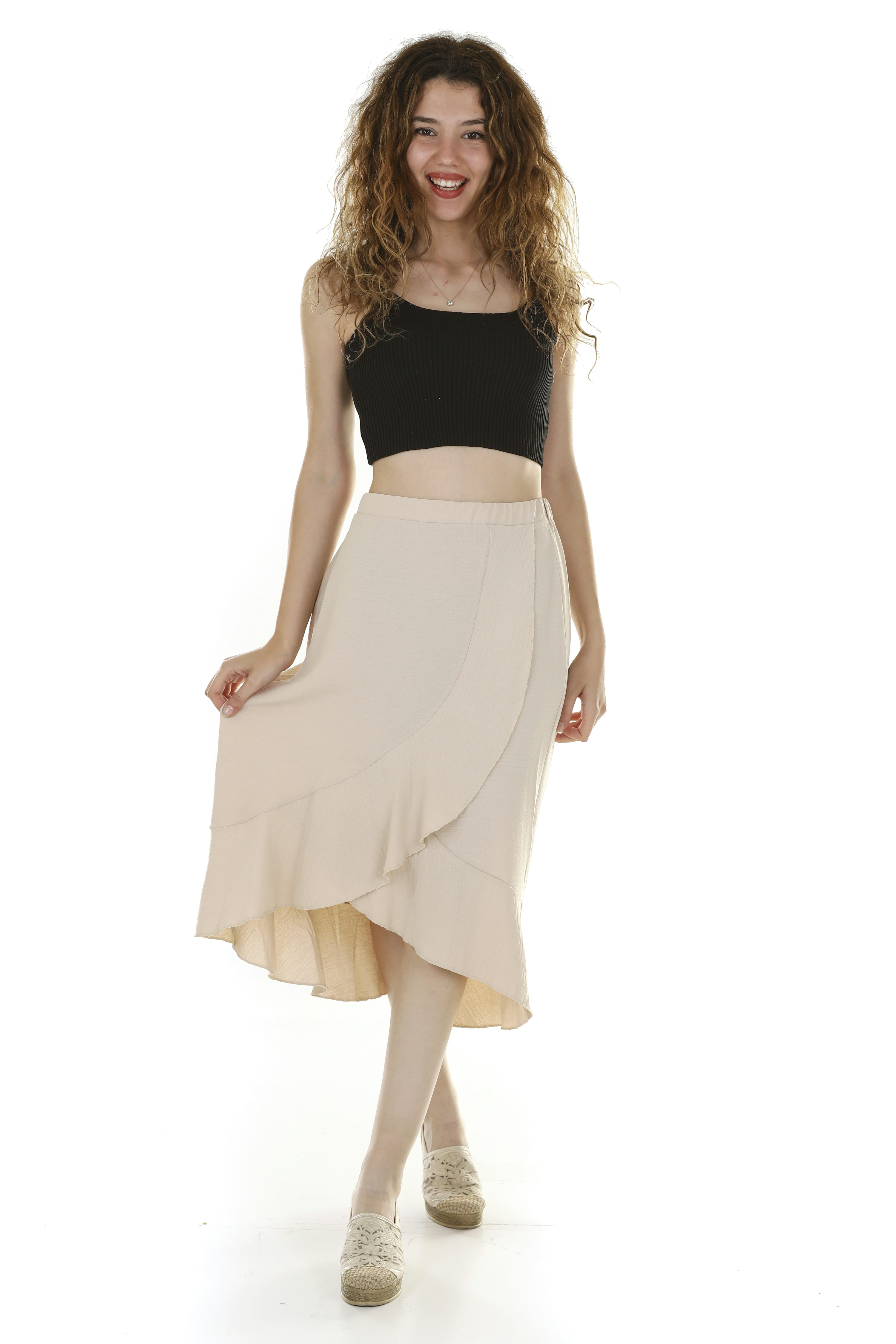 a young woman with curly hair is smiling and posing in a beige ruffle wrap skirt and a black crop top. she's wearing light beige espadrilles. the background is white, and the image is a studio shot. the overall style is casual and summery.