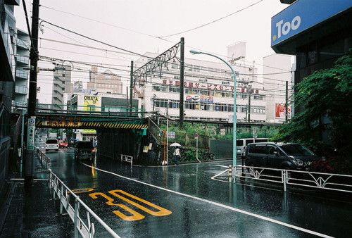 he image shows a rainy day scene on a city street in japan. the street is wet and reflects the buildings and surrounding environment. cars are parked and driving on the street. an overpass crosses above the street. buildings are visible in the background, and power lines and wires are strung across the sky. the overall atmosphere is gloomy and overcast. a speed limit sign is partially visible on the wet road.