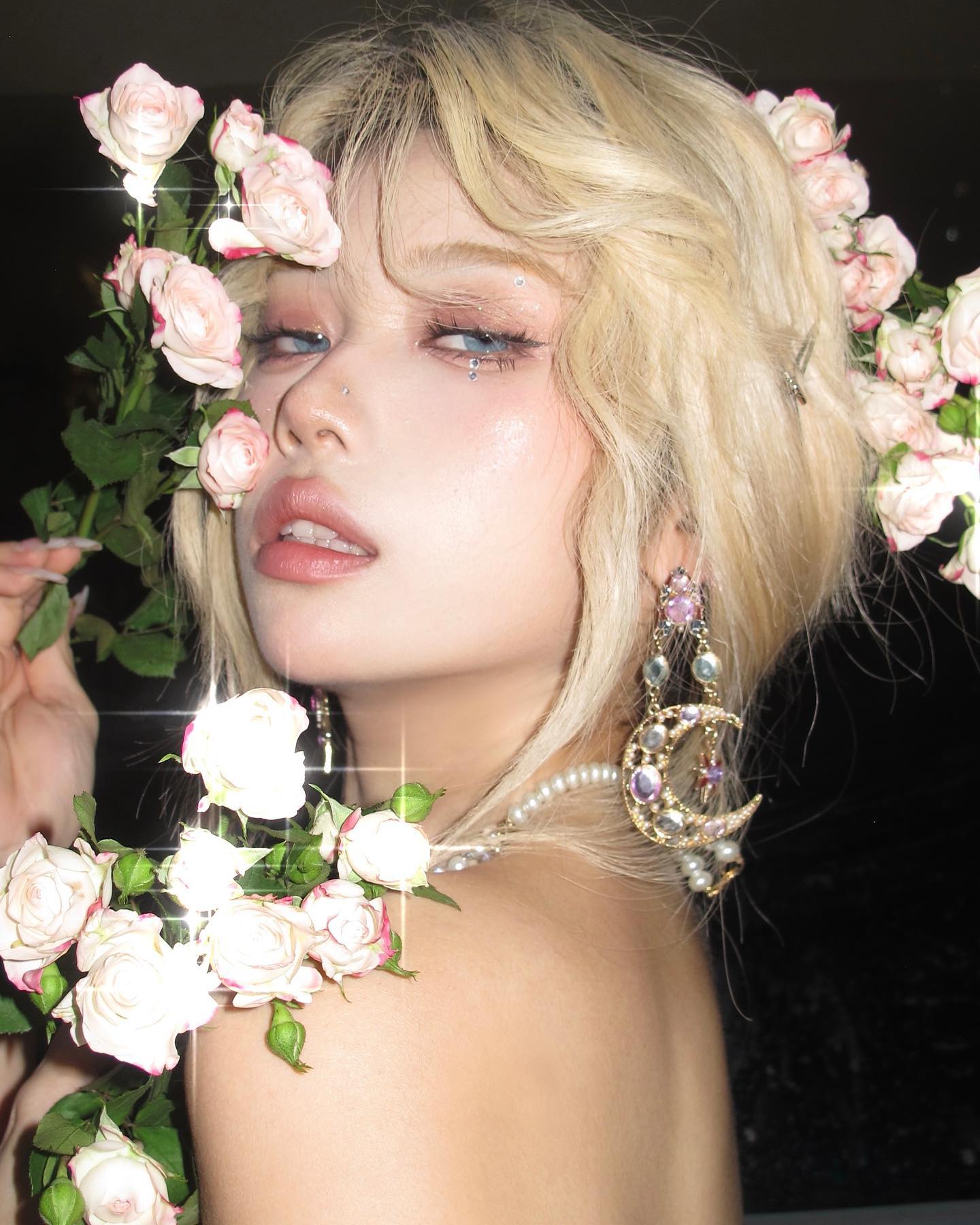 a closeup portrait of a young woman with blonde hair adorned with pink roses. she wears elaborate jewelry, including earrings with moon and star motifs, and has on shimmering eye makeup. the lighting is soft and flattering, highlighting her features and the delicate beauty of the flowers. her expression is alluring and slightly defiant. the overall aesthetic is feminine, trendy, and glamorous, with a hint of ethereal charm.