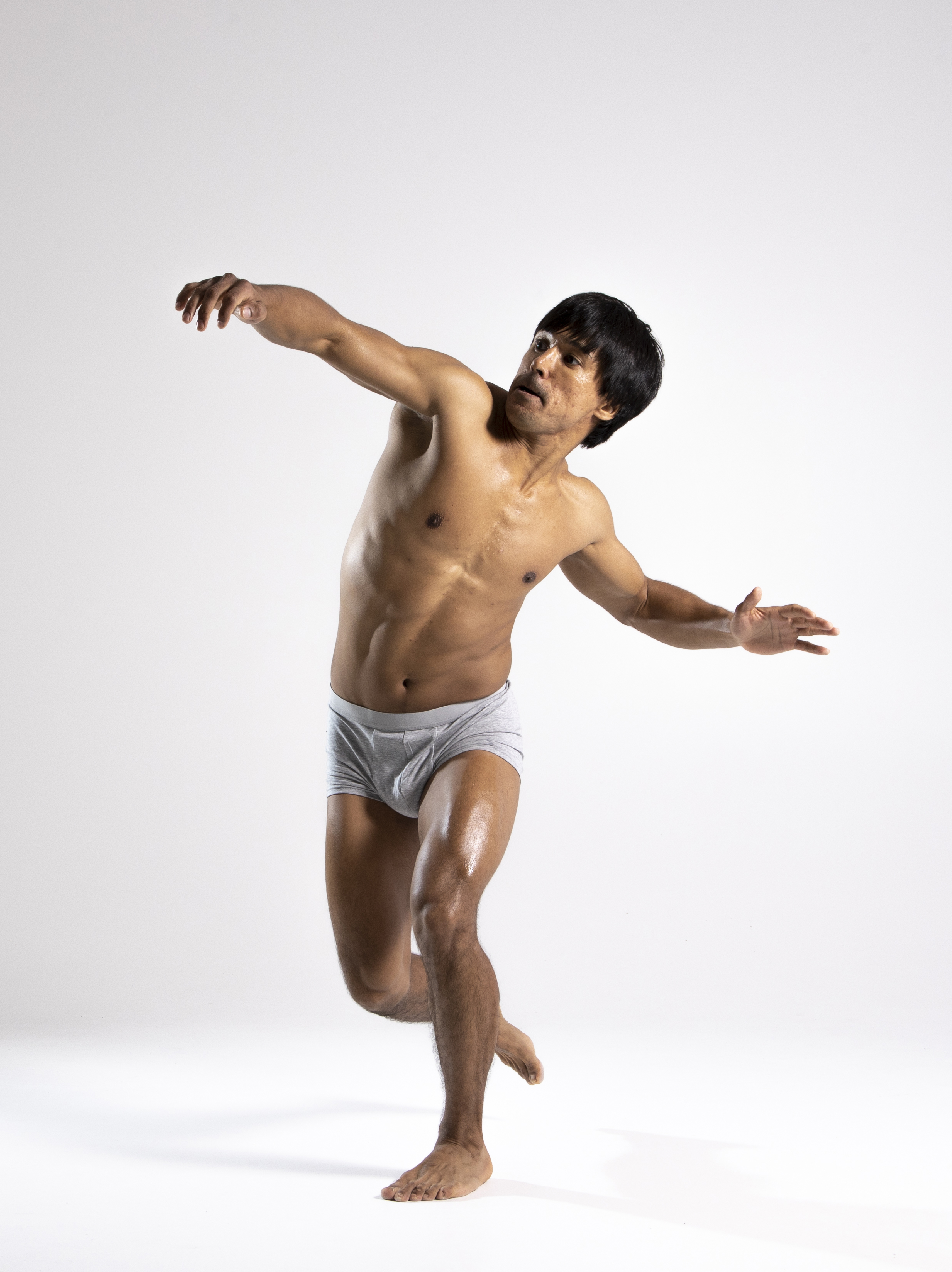 a shirtless man wearing underwear is dancing in a studio. he is facing the camera and has his arms outstretched. he is lit from the front and side.