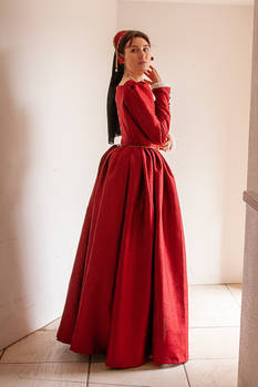 a woman in a long, elegant red gown stands in a room with white walls. the dress has long sleeves and a full skirt, typical of a historical style. the woman's hair is styled up, and she is wearing a red headpiece. her expression is somewhat serious. the overall appearance evokes a sense of historical fashion and elegance.