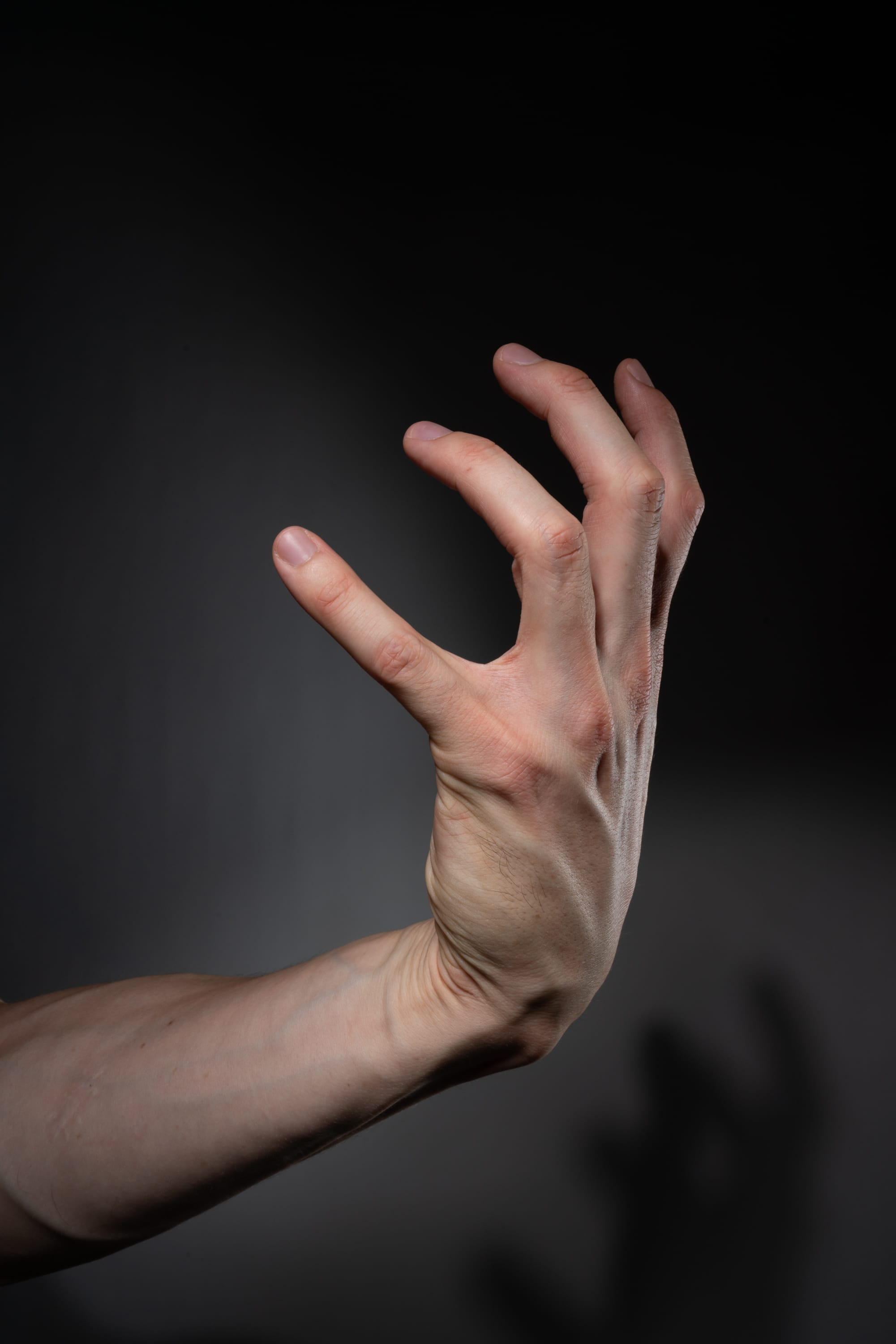 a man's left hand is reaching up with fingers spread against a dark grey backdrop, casting a dramatic shadow on the wall behind it. the studio lighting highlights the details of the skin and tendons, creating a sense of tension and anticipation.
