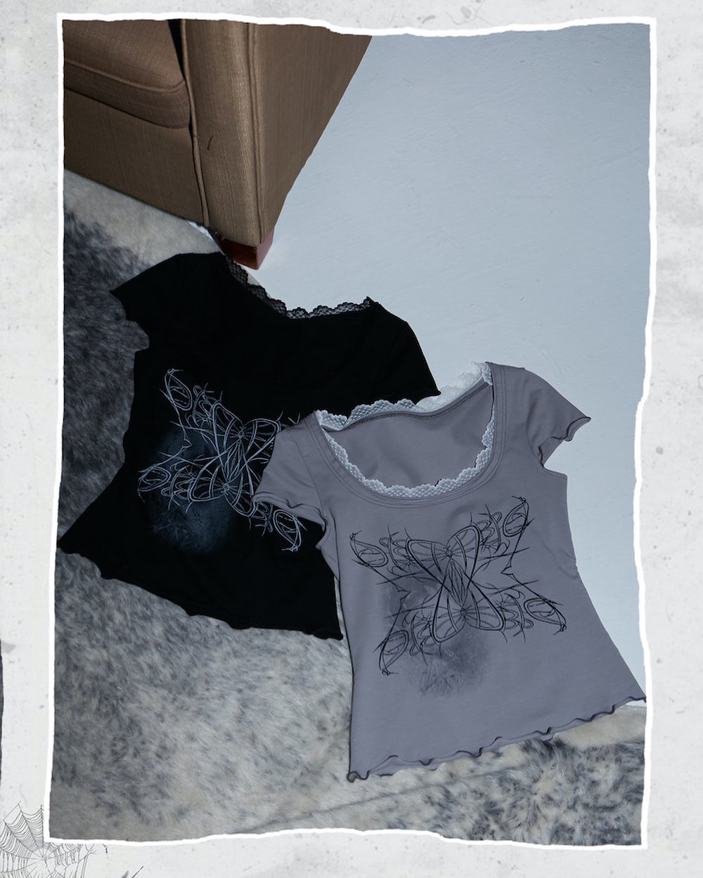 two shortsleeved tops lie on a faux fur rug in front of a brown couch. the top on the left is black, and the top on the right is grey. both tops are ribbed and have a lace trim around the neckline.  both tops feature a butterfly graphic.  the style is dark and edgy, possibly goth or grunge aesthetic. the overall image has a dark and moody feel.