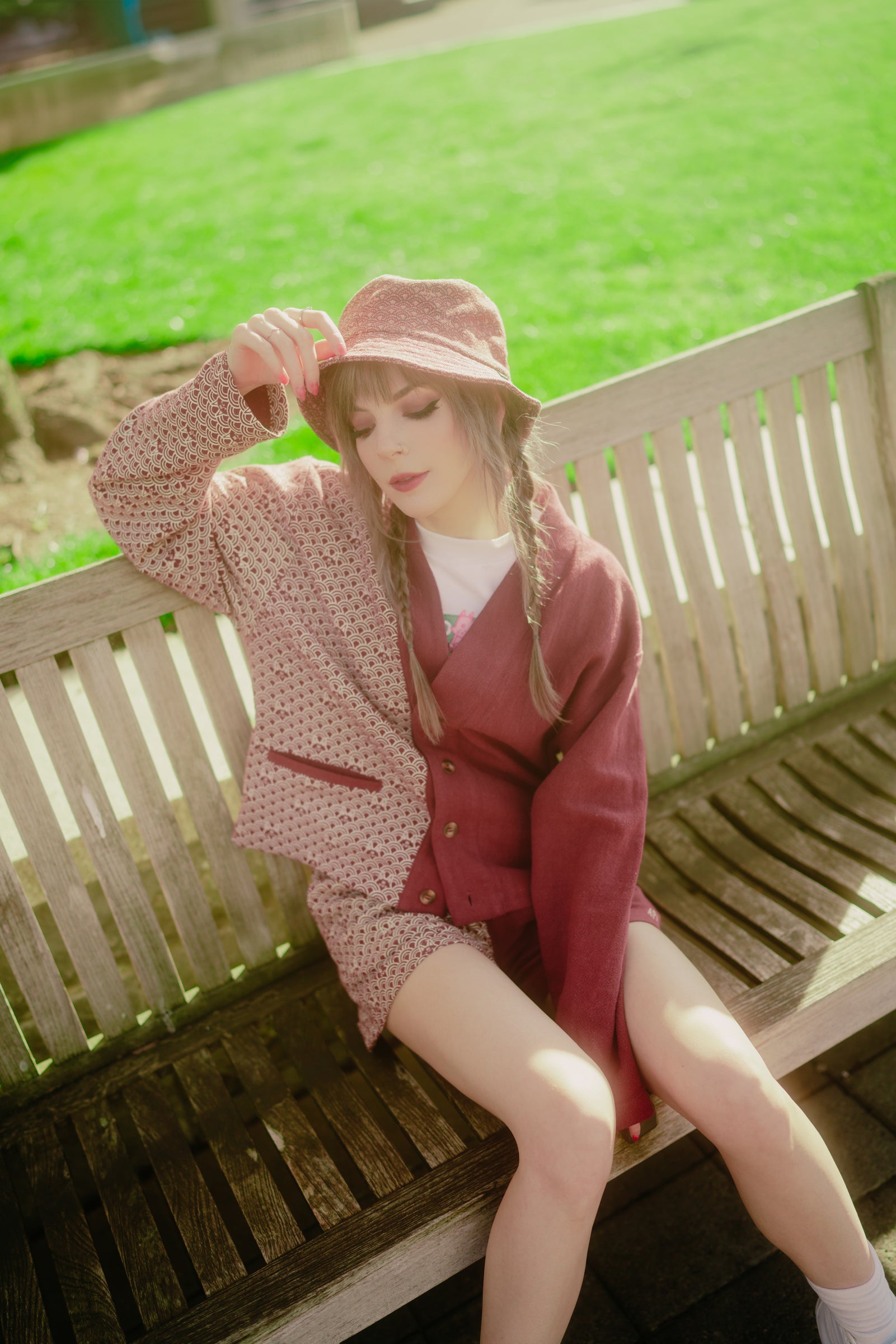a woman is sitting on a park bench wearing a pink and maroon splitdesign jacket and shorts with a matching bucket hat. she is fairskinned with blonde hair braided into pigtails. she is looking down while holding her hat. the background shows a green lawn and other park elements, suggesting an outdoor setting. the overall style is fashionable and trendy.