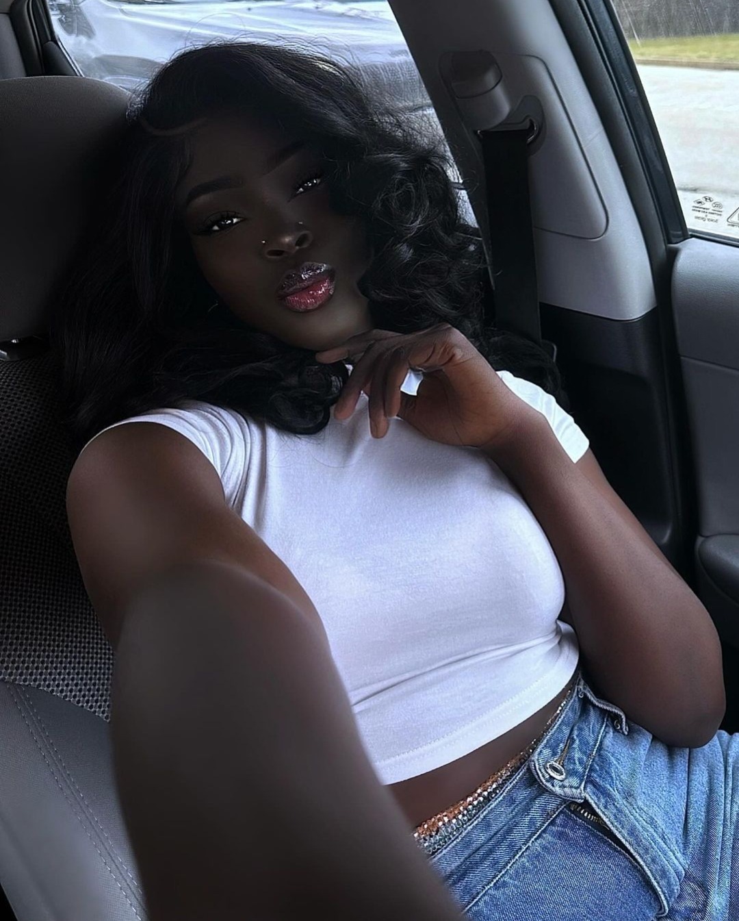 a young black woman with dark hair and stylish makeup takes a selfie from the passenger seat of a car. she is wearing a white tank top and blue jeans. the inside of the car is visible in the background.