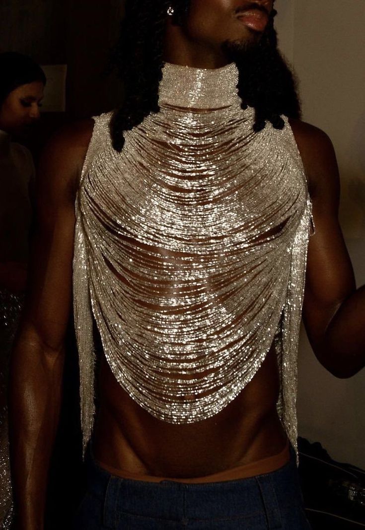 a close up portrait of a dark skinned man with long black hair, wearing a silver chain halter top that is sparkling, standing in front of a dark background. he is looking at the camera.