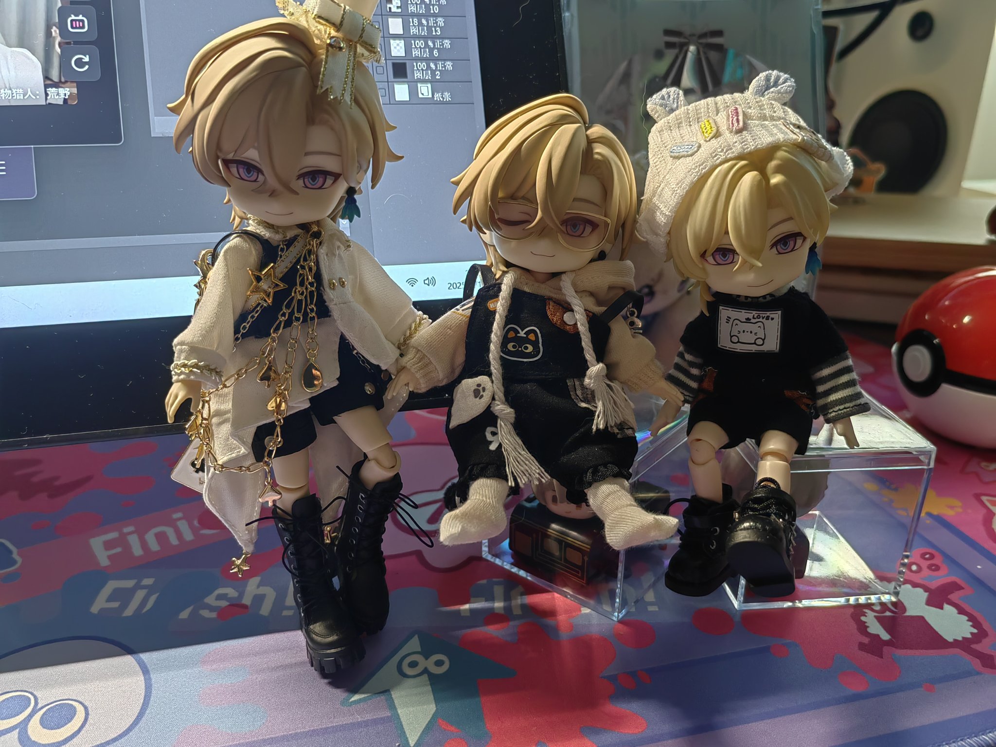 the image shows three animestyle dolls with blond hair and purple eyes. the dolls are dressed in different outfits: one has a white coat with gold chains and black boots, another is wearing overalls with a cat design and the third doll has a black shirt and a knit hat with ears. the dolls are posed against a backdrop that includes a computer screen, a pokemon ball, and a colorful desk mat. they appear to be collectible items.