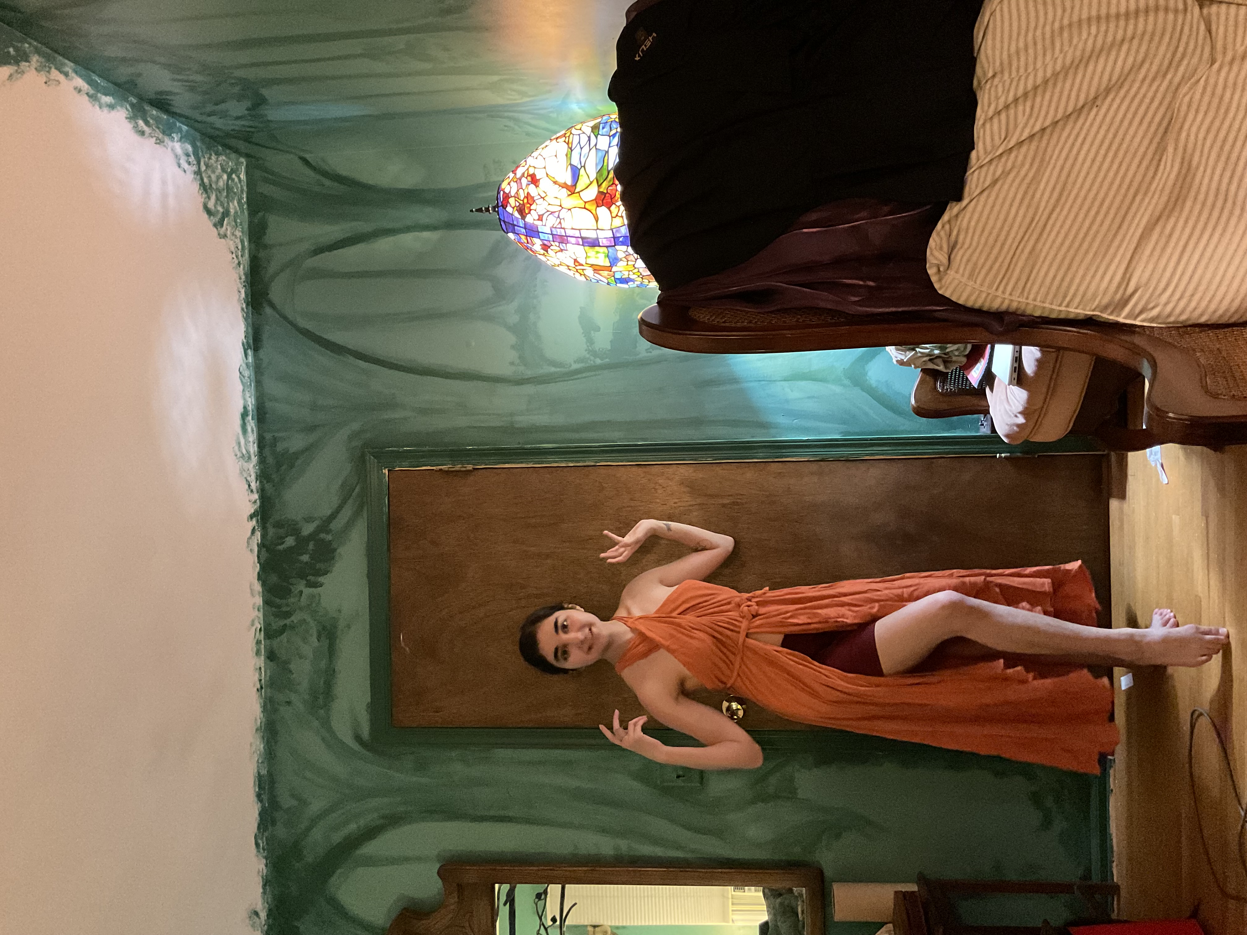 a young woman stands in a bedroom with distinctively painted green walls. she is wearing a flowing orange maxi dress with a halter neck and a high slit. the dress appears to be a wrap style.  the room features a vintagestyle armchair, a stainedglass lamp, and a mirror. the overall aesthetic is eclectic and slightly bohemian.  the young woman is posing for a photo, hands near her waist.