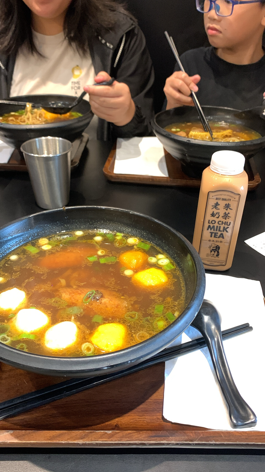 photo taken from above shows two people enjoying a meal of asian noodle soup. the foreground features a large bowl of broth filled with meatballs, vegetables, and noodles. black chopsticks rest on the bowl and a brown placemat.  two other bowls of similar soup can be seen behind the first, accompanied by metal chopsticks and a metal cup.  the individuals are partially visible, the adult holding a spoon and the child holding chopsticks. a bottle of "lo chu milk tea" sits on the table next to the bowls.  the overall color palette is warm and muted.