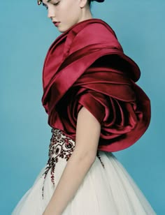 a young woman in a red and white gown stands against a blue background. she has her hair up in an elegant updo and is wearing red fabric around her shoulders, which looks like a large flower or rose. she is looking off to the side, with a thoughtful expression on her face. the dress is elegant and flowing, and the woman's pose is graceful. the image is a beautiful and timeless portrait of a woman in a highfashion gown.