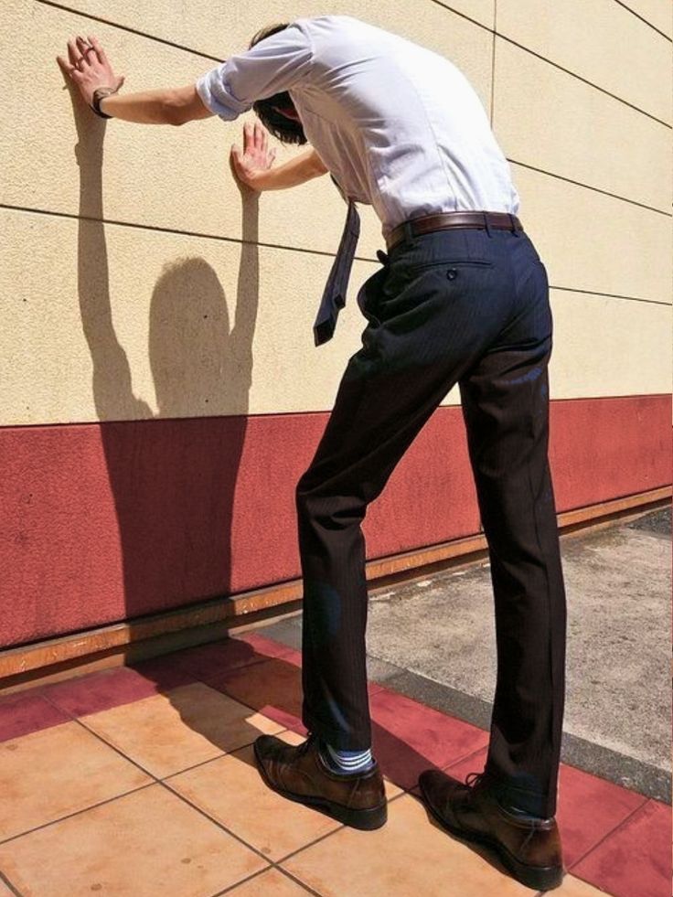 a man in a white shirt and dress pants is leaning against a wall with his arms and head resting on the wall.  the sunlight casts his shadow onto the wall. he appears to be exhausted and frustrated, possibly from work stress.  he is wearing a blue tie and brown dress shoes with white socks.