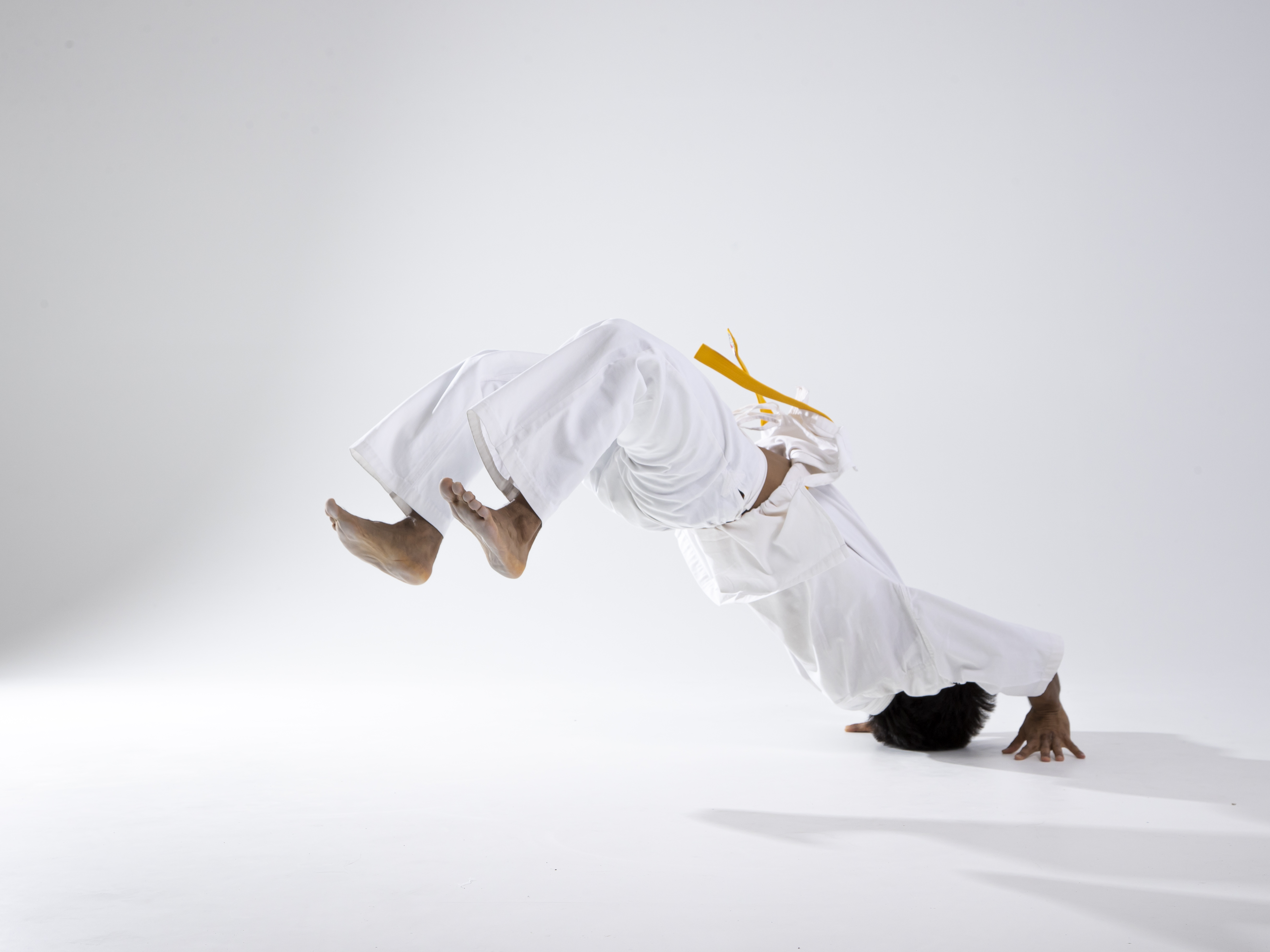 a man in a white judo gi and yellow belt is performing an acrobatic tumble in a studio. he is upside down with his head and hands touching the floor, and his feet in the air. he is against a white background.