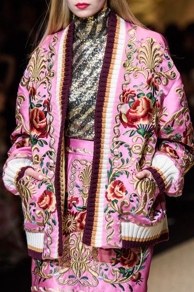 loseup view of a female model showcasing a pink embroidered jacket. the jacket features intricate floral patterns with roses and gold detailing, a maroon and white ribbed trim, and is worn over a gold sequined turtleneck top and matching pink skirt. the overall style is luxurious and elegant. the image is likely from a fashion show or photoshoot.
