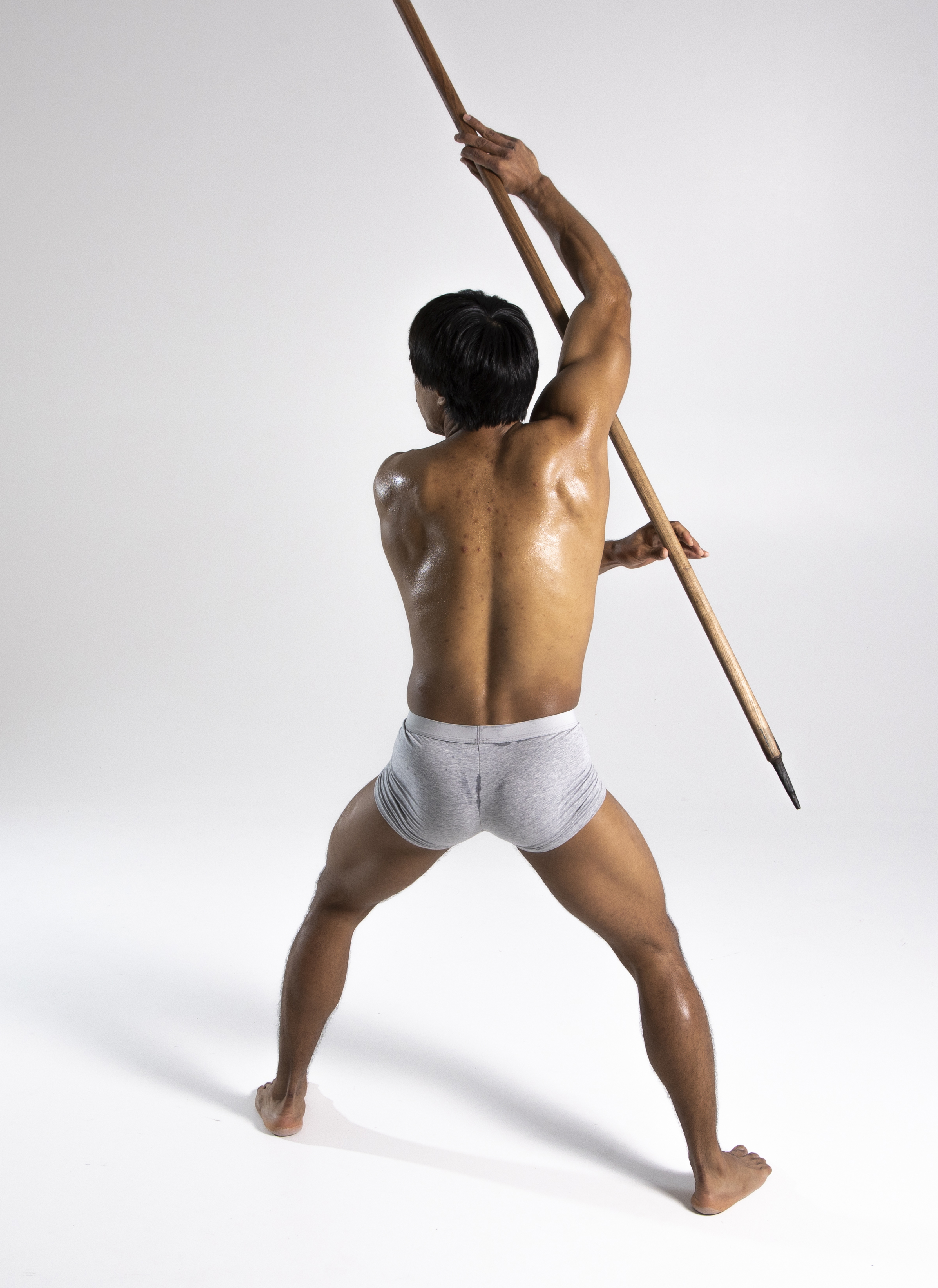 a shirtless, muscular man wearing a loincloth stands with bare feet in a studio shot. he is seen from the back, holding a spear high above his head. the image is a full shot, emphasizing his physical build and the weapon he wields.