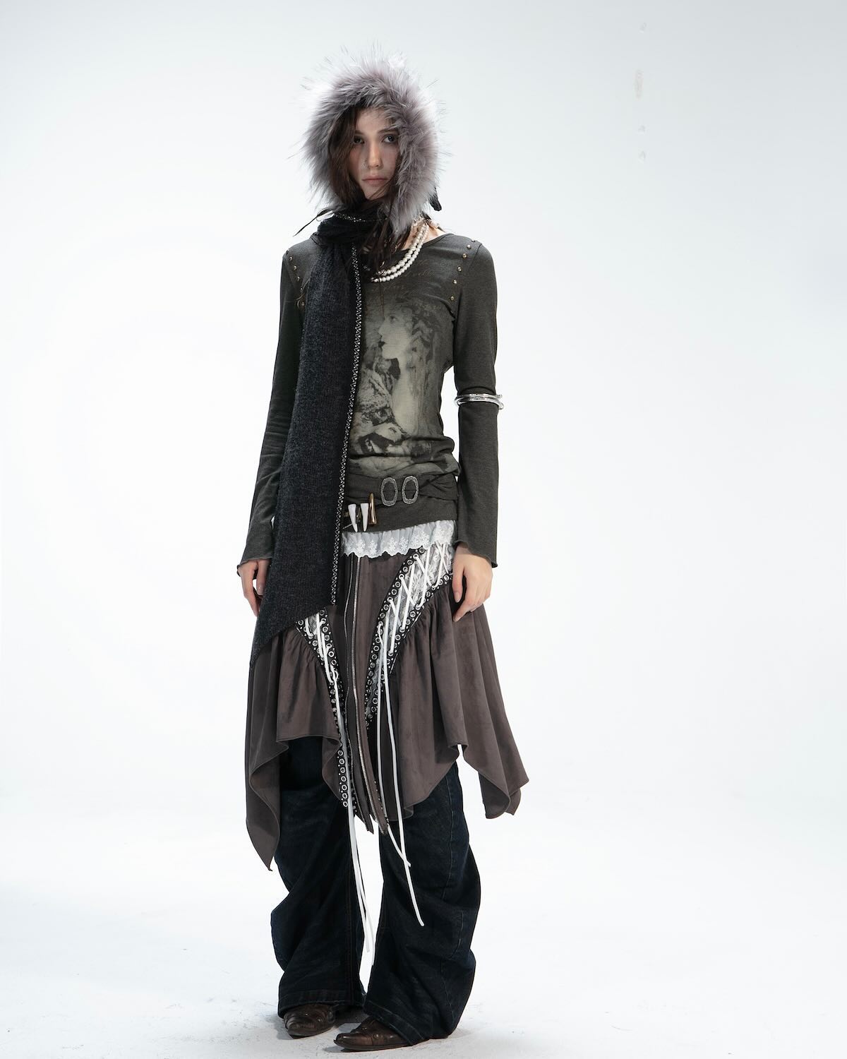 fullbody shot of a female model showcasing a stylish outfit. the model is standing against a white background, wearing a longsleeved dark gray top featuring a graphic print, a gray skirt with lace detailing and a unique asymmetrical design. she accessorizes with a dark gray hooded fur scarf and a belt. the model's jeans are visible from below the skirt. the overall aesthetic is a mix of grunge, bohemian, and chic elements, creating a fashionable winter or fall look.