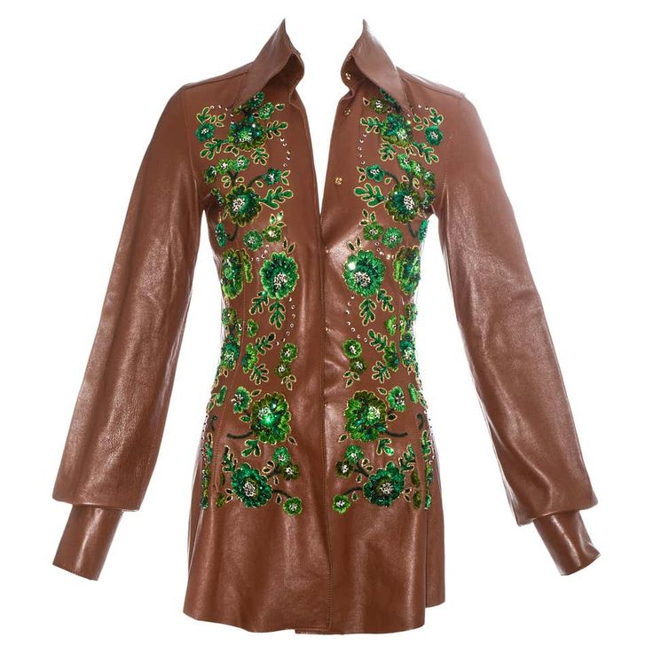 a brown leather jacket with a green floral pattern and beads. the jacket has long sleeves and a collared neckline. the jacket is fitted and has a slightly flared hem. the fabric of the jacket appears to be leather, and it has a glossy finish.