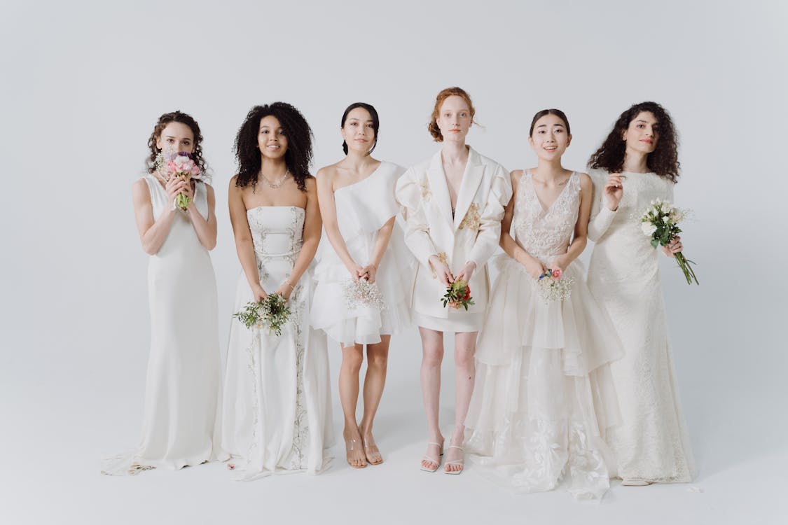 a photograph shows six diverse women dressed in various white bridal gowns, standing in a row against a white background. each woman holds a small bouquet of flowers.  the dresses are all different styles – some are simple, others are more ornate and layered. the women are smiling, suggesting a happy and celebratory atmosphere, likely a bridal party photoshoot.