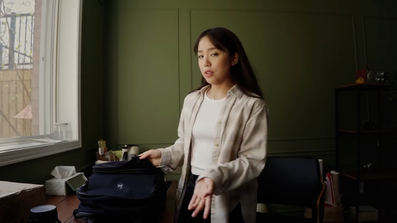 a young woman is standing in a room with sunlight streaming in from a window to her left. she's wearing a white tshirt and an unbuttoned light beige plaid shirt.  she has a black backpack open on the desk in front of her and appears to be talking to the camera, potentially about the contents of her bag. the walls of the room are a deep green and there is a wooden shelf to her right.