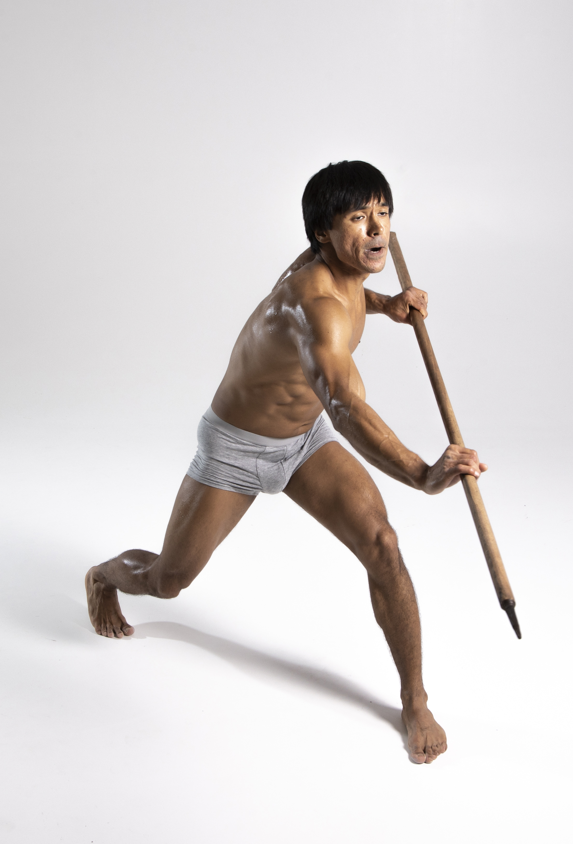 a muscular asian man is posed in a fulllength studio shot. he is barechested, wearing only gray underwear, and holding a long wooden spear. he is barefoot and appears to be in a dynamic, athletic stance.