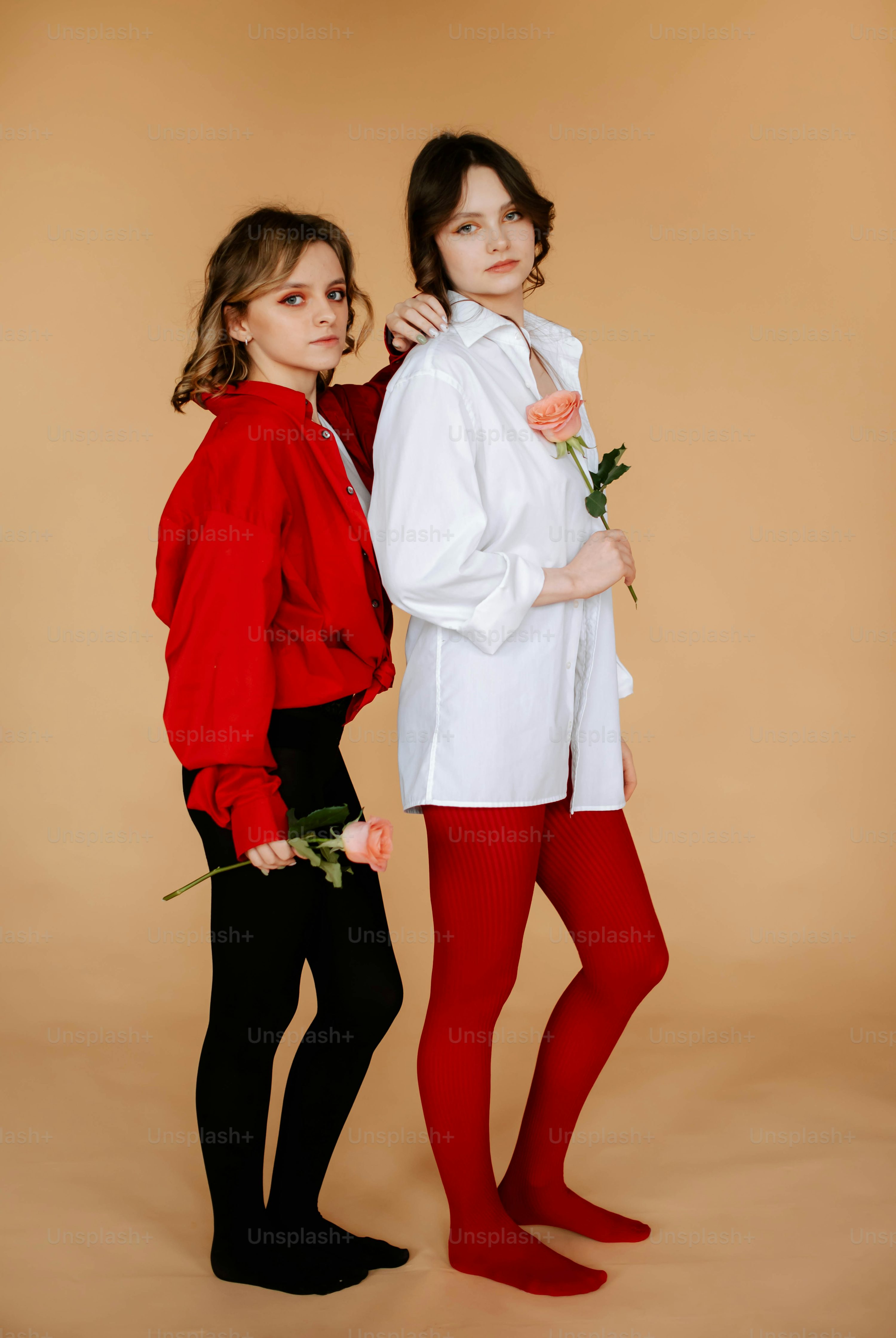 two young women are posing together in a studio shot against a beige background. one girl is wearing a red jacket and black tights, while the other girl is wearing a white shirt and red tights.  both girls are holding a single rose. they appear to be friends, and the image has a stylish and trendy feel.