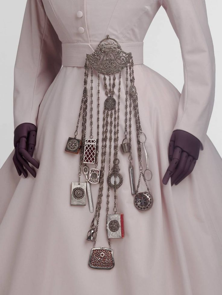 the image shows a light pink dress on a mannequin, with a wide belt adorned with elaborate silver jewelry and accessories hanging from it. the jewelry includes various pendants, charms, and a small purselike ornament attached to chains. the mannequin has dark purple hands.