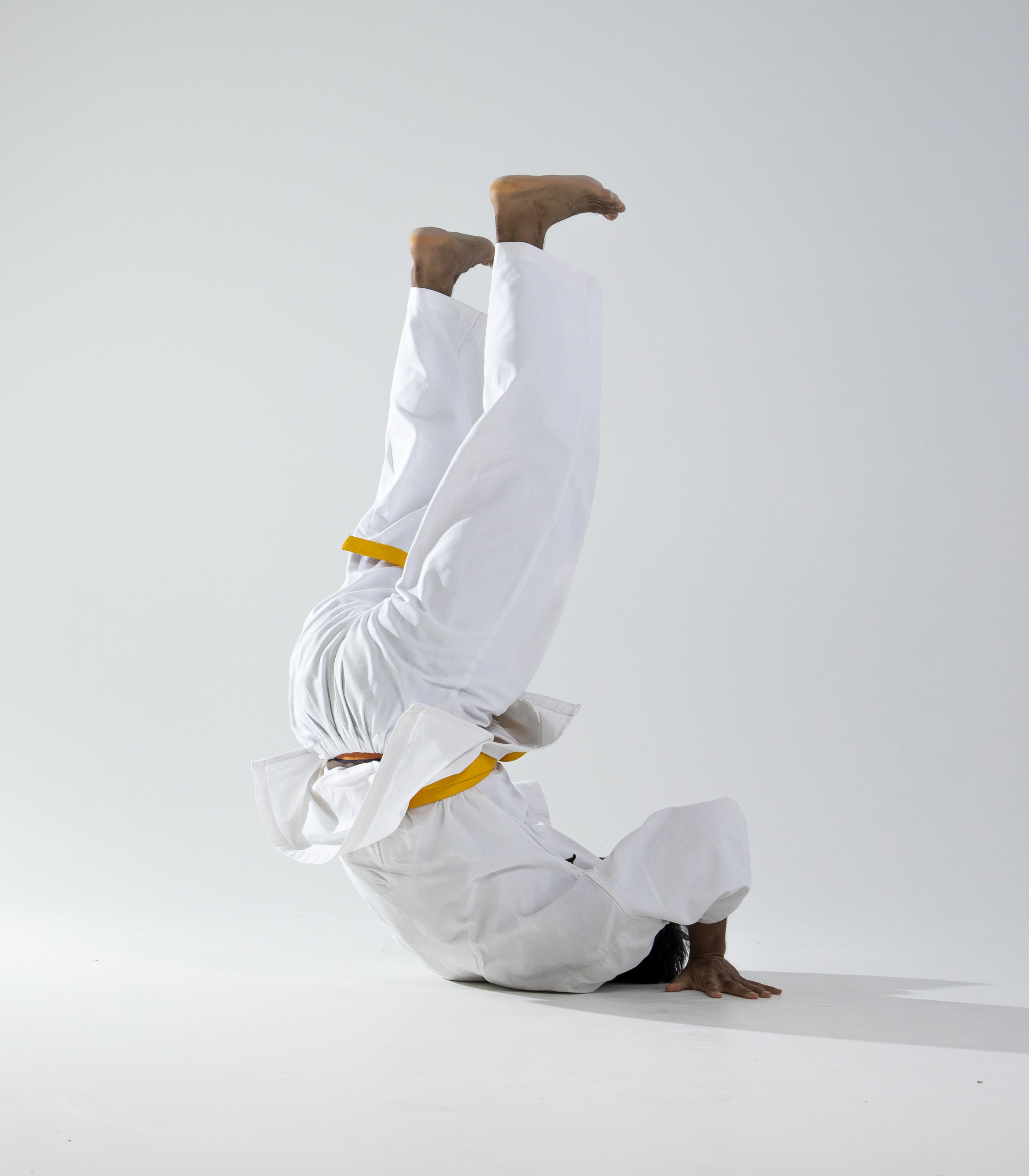 a man wearing a white martial arts uniform with a yellow belt is upside down, balancing on his hands in a martial arts pose. his legs are straight up in the air and his feet are bare. he is practicing martial arts and demonstrating balance and control. the background is white.