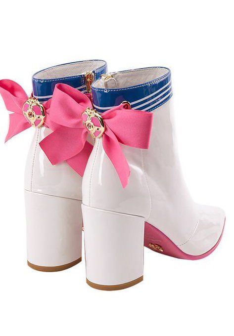 a pair of white, patent leather ankle boots with chunky heels are displayed on a white background. each boot features a pink ribbon with a gold heartshaped buckle on the back. the top of each boot has a blue stripe design reminiscent of a sailor uniform. the sole of the right boot is visible and is pink.