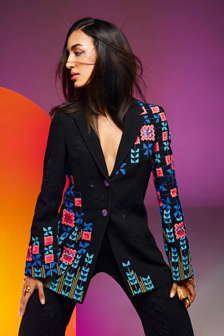 a woman is standing, wearing a black blazer with floral embroidery and purple buttons. the blazer is open, revealing a glimpse of skin underneath. she is adorned with rings and is looking away from the camera. the background features a colorful gradient.