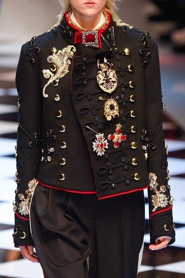a woman is wearing a black militarystyle jacket with elaborate gold and red embellishments. the jacket features numerous gold buttons, ornamental details on the shoulders and sleeves, and several decorative brooches pinned to the front. a red trim runs along the edges of the jacket. she is also wearing black pants. the outfit is presented in a highfashion context.