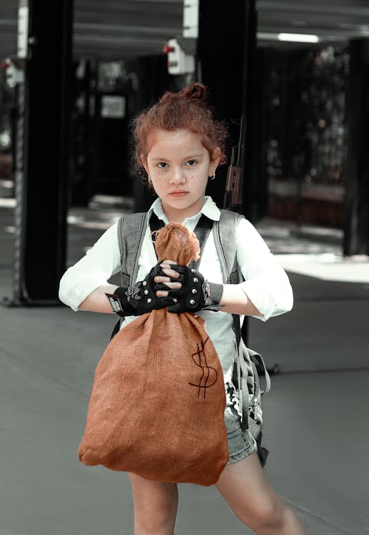 a fullbody shot of a young redhead girl, seriously posing with a burlap money bag in her hands. she is wearing a white shirt, denim shorts, a dark vest, and fingerless gloves. the background is a muted urban alleyway. the image has a slightly desaturated, gritty, and film noir style with selective color emphasizing the girl and money bag. the overall impression is one of a tough, young female character.