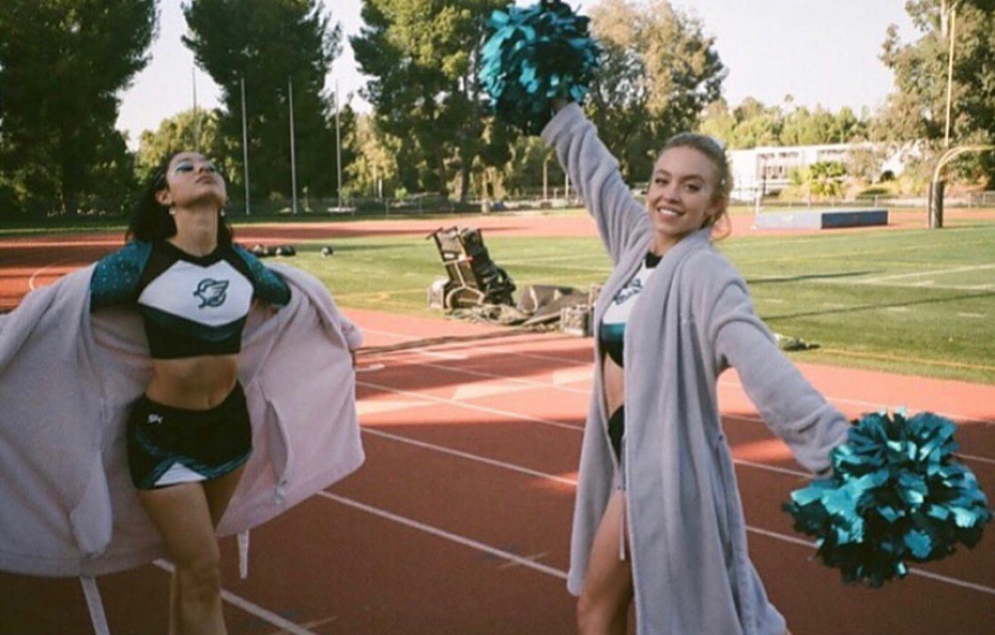 the image shows two teenage girls on a school running track. one girl is wearing a teal and white cheerleading uniform and is holding a light lavender colored bathrobe open around her.  the other girl is wearing a light grey bathrobe and holding a teal pompom; she is smiling at the camera.  both girls have teal and white pompoms. the background is a grassy field with trees and a school building visible in the distance.  the overall atmosphere is relaxed and cheerful, suggesting a break or informal moment between cheerleading activities.