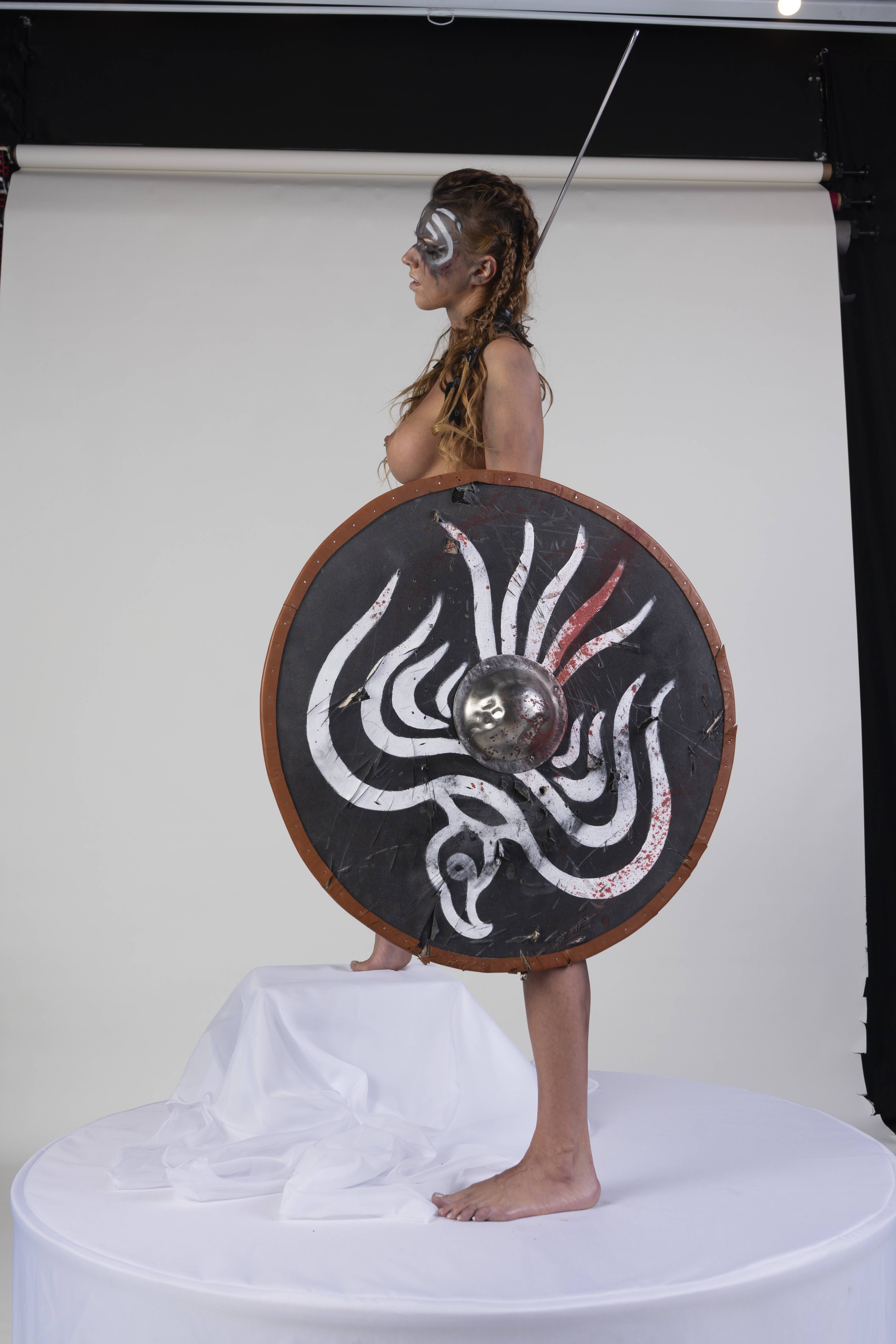 a partially nude woman with makeup and braids, wearing a headdress and holding a shield and sword, stands on a platform. she is barefoot and looking away.