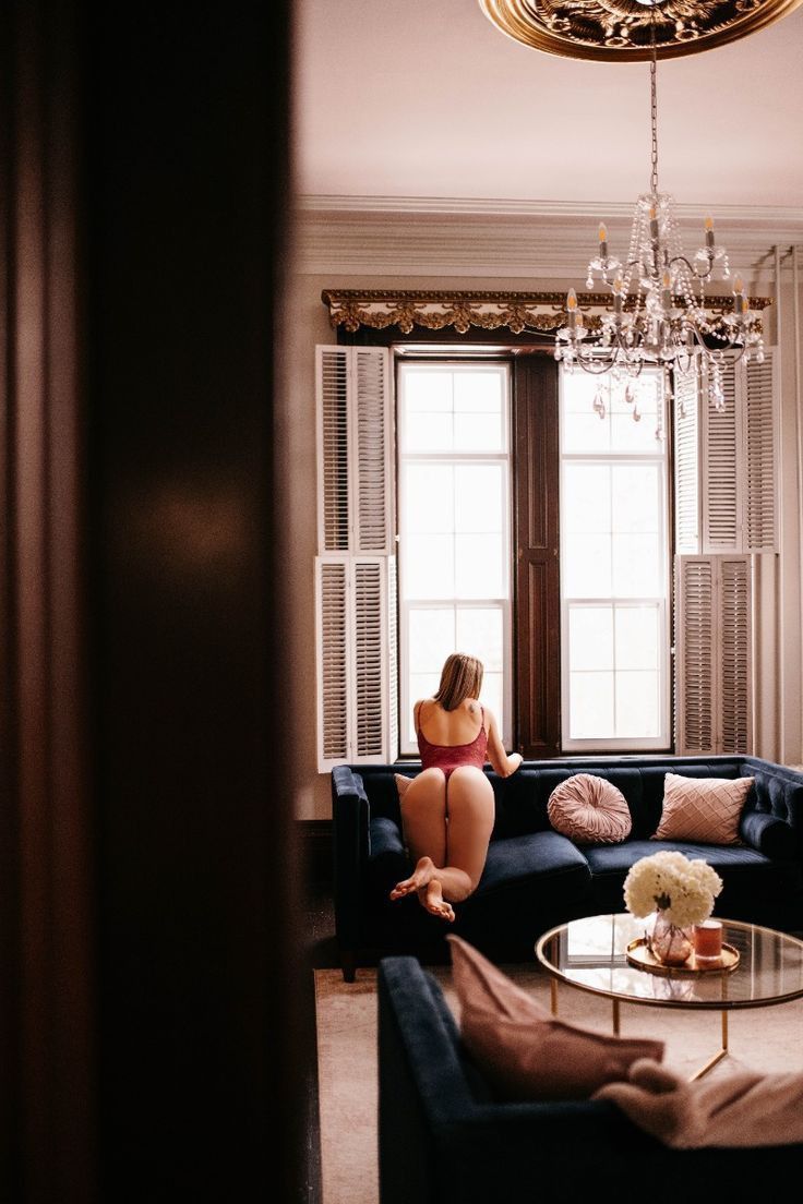 a woman in a red bodysuit is sitting on a dark blue couch, gazing out a large window with white shutters. the room is elegantly decorated with a crystal chandelier, ornate ceiling, and luxurious furniture. the overall mood is sensual and intimate. the photo is taken from a slightly obscured angle, adding to the feeling of privacy. the image also includes a coffee table with flowers and other decor.