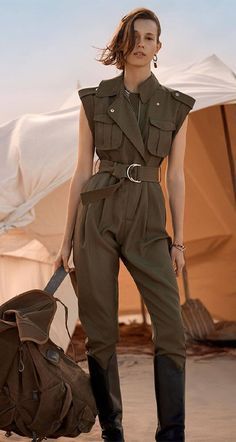 a woman in a stylish green jumpsuit, complete with a belt and a brown leather backpack, stands confidently in a desert setting. she has short, styled hair, makeup, and is accessorized with earrings and sunglasses. the warm desert colors create a sense of adventure and stylish travel.