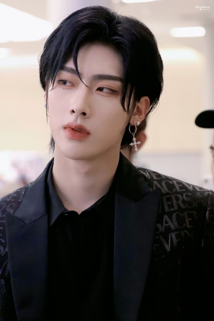this is a picture of a young man with styled black hair and makeup. he is wearing an earring in his left ear and a black jacket over a black shirt. he is looking to the left of the camera. the background is blurred, but it appears to be indoors and welllit.