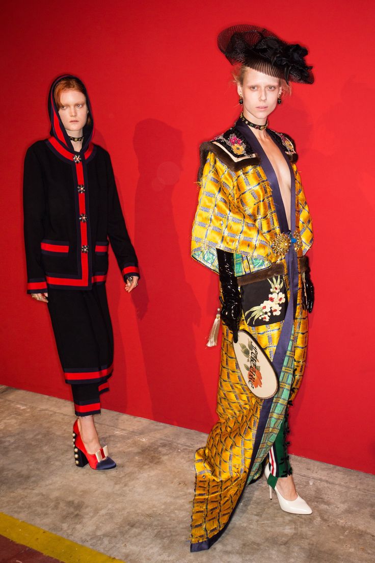 two models are posing against a red backdrop. the model on the left is wearing a navy blue coat with red and white trim and red and blue shoes. the model on the right is wearing a long yellow kimonostyle robe with an embroidered design, black gloves, a black hat, and white heels.  both outfits appear to be part of a gucci collection. the image is likely taken backstage at a fashion show.