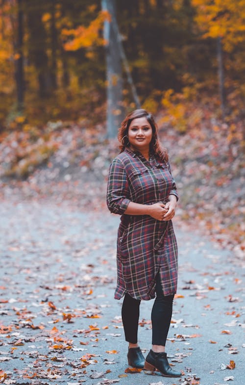 the image shows a smiling young woman of south asian descent standing on a pathway in a park or wooded area during autumn.  the ground is covered in fallen orange and brown leaves. she is wearing a longsleeved plaid shirt dress over black leggings and black ankle boots. the background is softly blurred, focusing attention on the woman. the overall mood is warm, inviting and pleasant, suggesting a feeling of autumnal comfort.