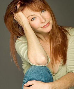 a woman with long red hair is sitting in a studio setting. she is wearing a green striped shirt and blue jeans. her hair is parted on the side and she is smiling at the camera. her right arm is resting on her head.