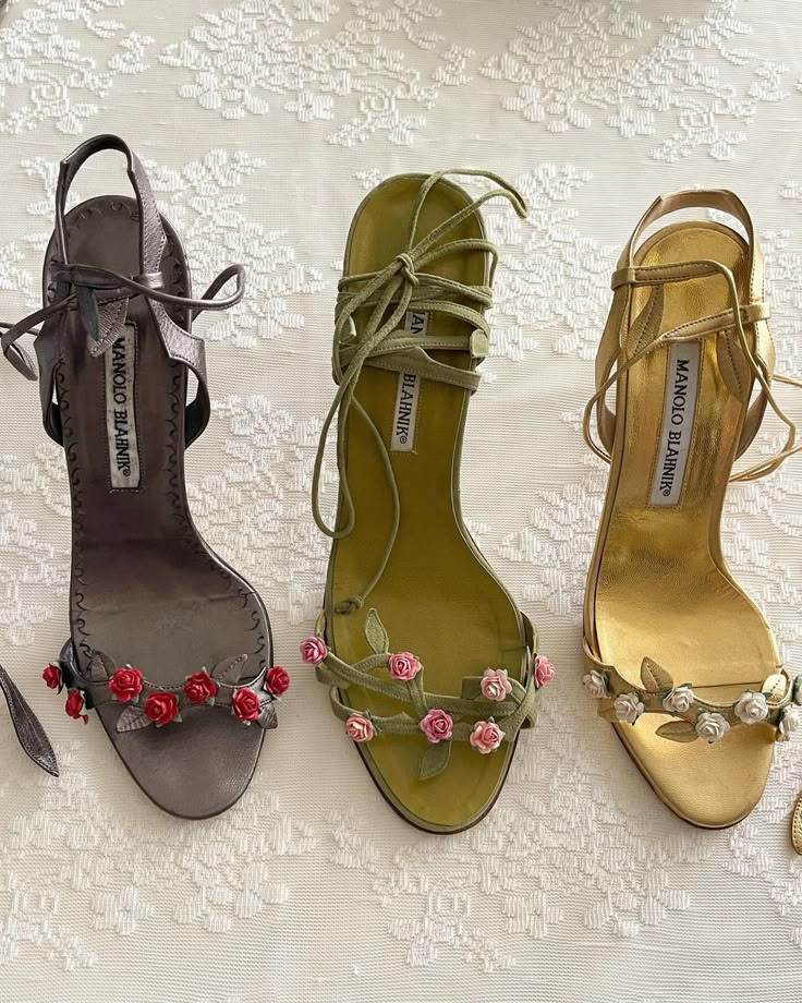 three pairs of manolo blahnik heeled sandals are arranged in a row on a patterned white surface. the sandals feature flower embellishments along the front strap. the leftmost pair is grey with red roses, the middle pair is green with pink roses, and the rightmost pair is gold with white roses. each sandal has delicate straps and the manolo blahnik label is visible inside.