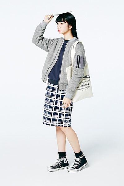 a young asian woman stands casually in a studio. she's wearing a gray bomber jacket, a black plaid skirt, a canvas tote bag, and black hightop converse sneakers.  she is holding a piece of paper over her head with her hand.