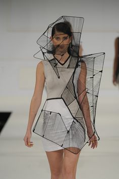 a female model poses on a runway wearing a futuristic, geometric black and white dress. the dress has a unique design that incorporates wire mesh and is draped over the body in a way that is both stylish and elegant.