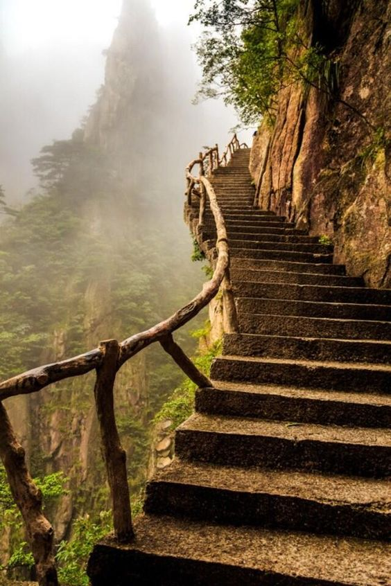 the image shows a steep stone staircase with a wooden railing that ascends a mountainside shrouded in fog. lush greenery clings to the surrounding cliffs, creating a sense of natural beauty and a challenging climb. the stairs lead up into the misty heights, evoking a feeling of mystery and adventure.