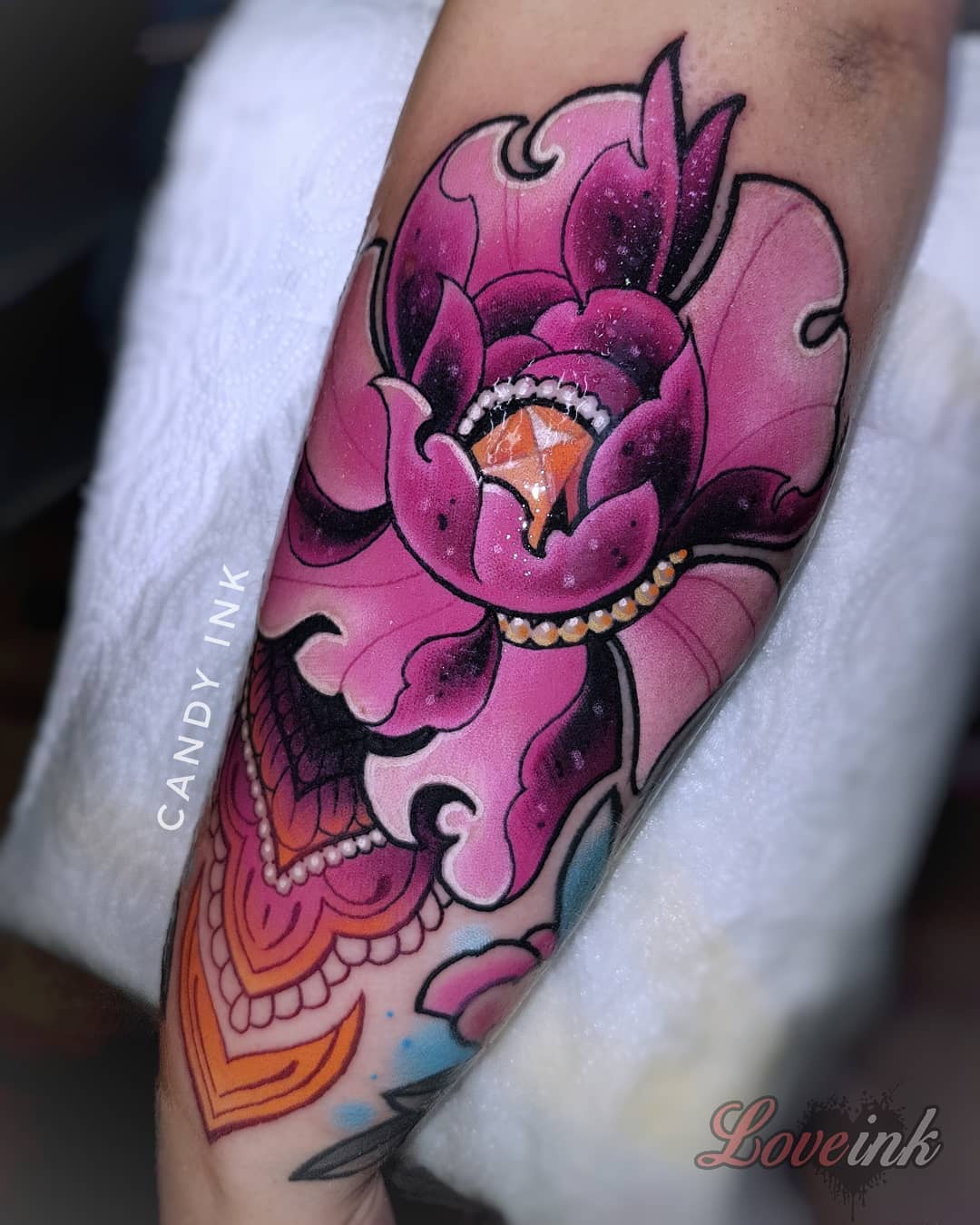 a closeup photograph showcasing a vibrant neotraditional tattoo of a peony flower on an individual's forearm. the tattoo, rendered in a striking palette of pinks, purples, and oranges, features a large peony blossom as its centerpiece. a sparkling jewel adorns the heart of the flower, adding a touch of opulence. the blossom is further enhanced by delicate white accents that mimic the effect of glitter, lending the design a luminous quality. below the peony, the tattoo transitions into an intricate pattern of orange and purple, creating a sense of visual flow and echoing the organic forms of the flower above. the tattoo is credited to "candy ink" and features a watermark for "love ink," suggesting either the artist's or the studio's name. the photograph is taken indoors under bright lighting, allowing the colors of the tattoo to pop against the individual's skin. the closeup perspective draws the viewer's attention to the intricate details of the tattoo, highlighting the artist's skill in blending colors and creating a sense of depth and dimension.