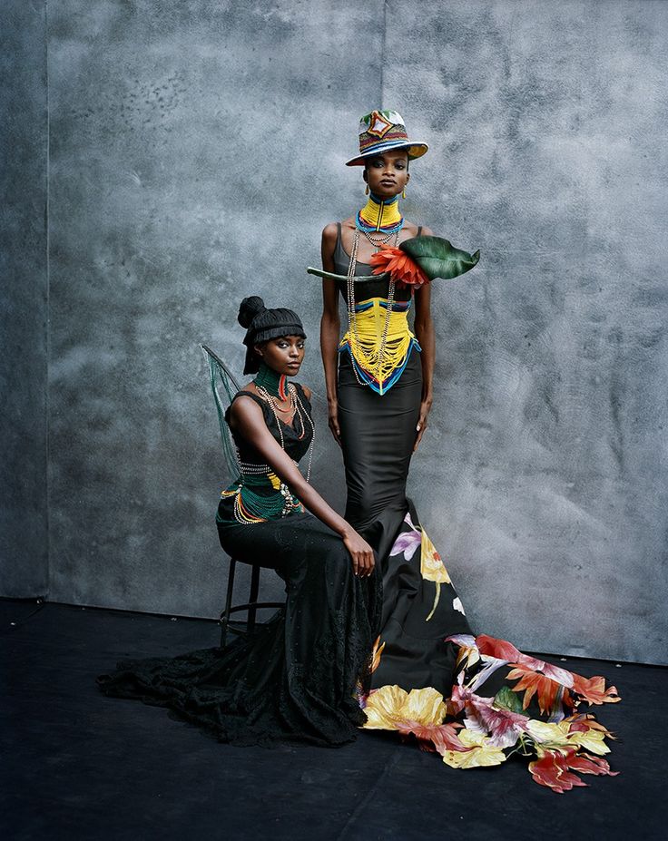 two black models are posing in fashion. the model on the left is sitting on a stool while the model on the right is standing. both models are wearing black dresses with floral accents, hats, and jewelry. the model on the left has a green and black dress, while the model on the right has a yellow and black dress. the background is a gray wall.