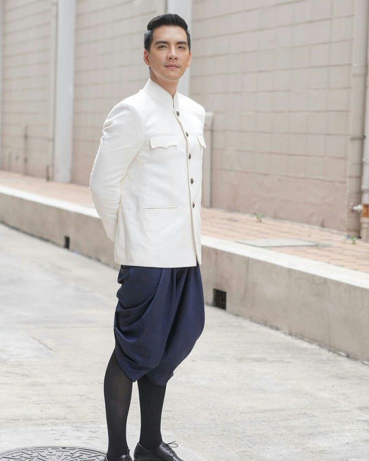a man in a white jacket, blue pants, and black shoes standing in front of a white wall. he is looking at the camera with a smile. he is dressed in a traditional thai suit.