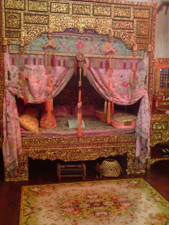 a color photograph shows an ornately decorated canopy bed with an elaborate headboard and footboard, featuring gold accents, pink floral curtains, and a floral rug in front. the bed is adorned with multiple pillows, and the background suggests it may be part of a display. a small woven basket sits beneath the bed.